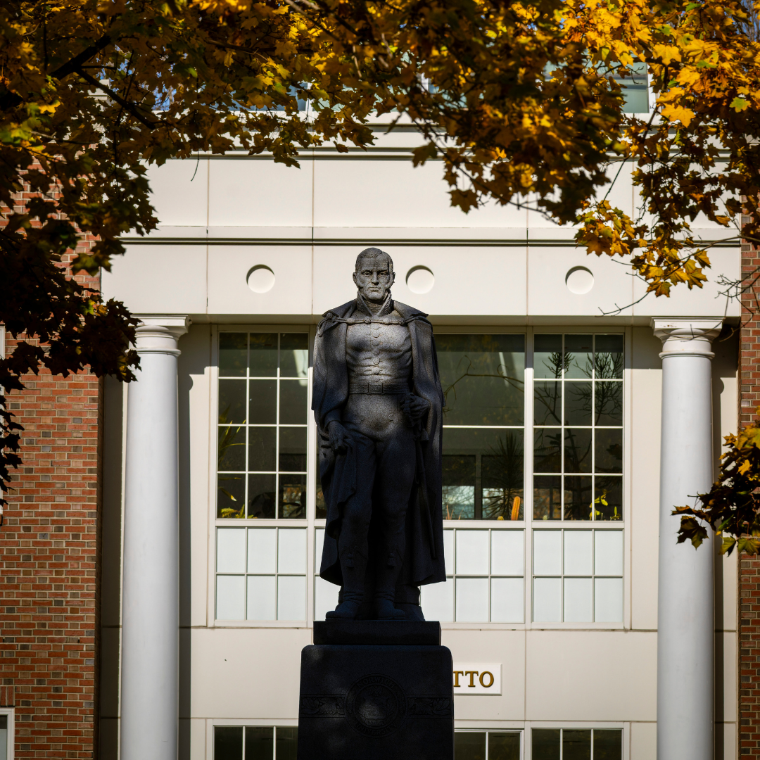 A statue of Alden Partridge stands in the foreground framed by autumn-colored leaves, with a classic building featuring white columns in the background.