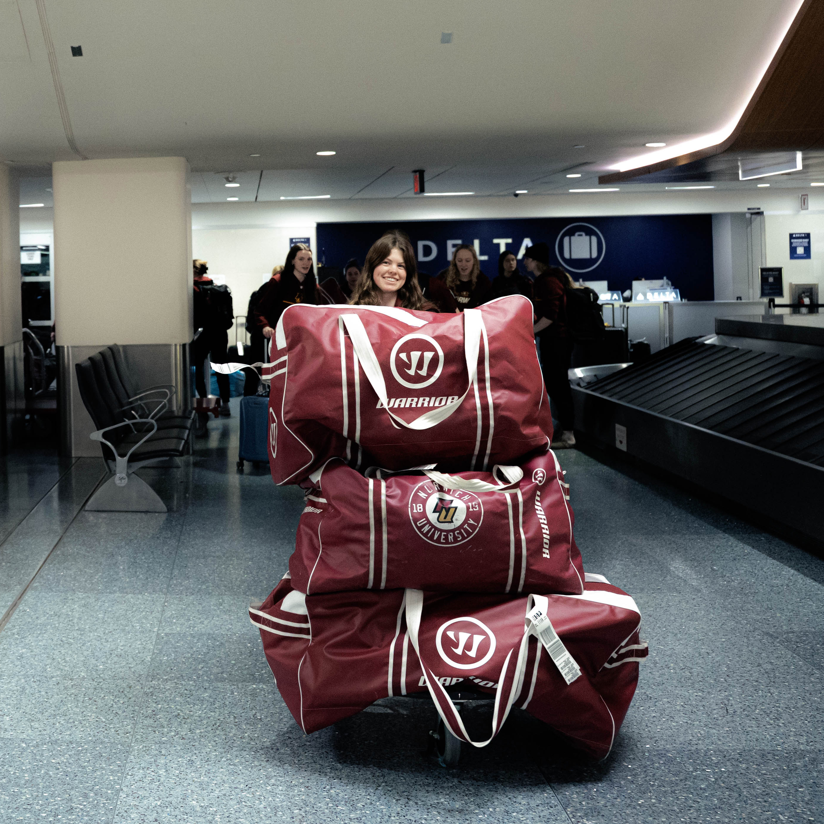 Student pushes cart stacked with Norwich University duffel bags through an airport baggage claim area.