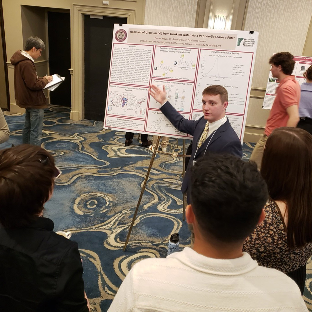 Student presents a research poster to a group at an indoor academic conference.