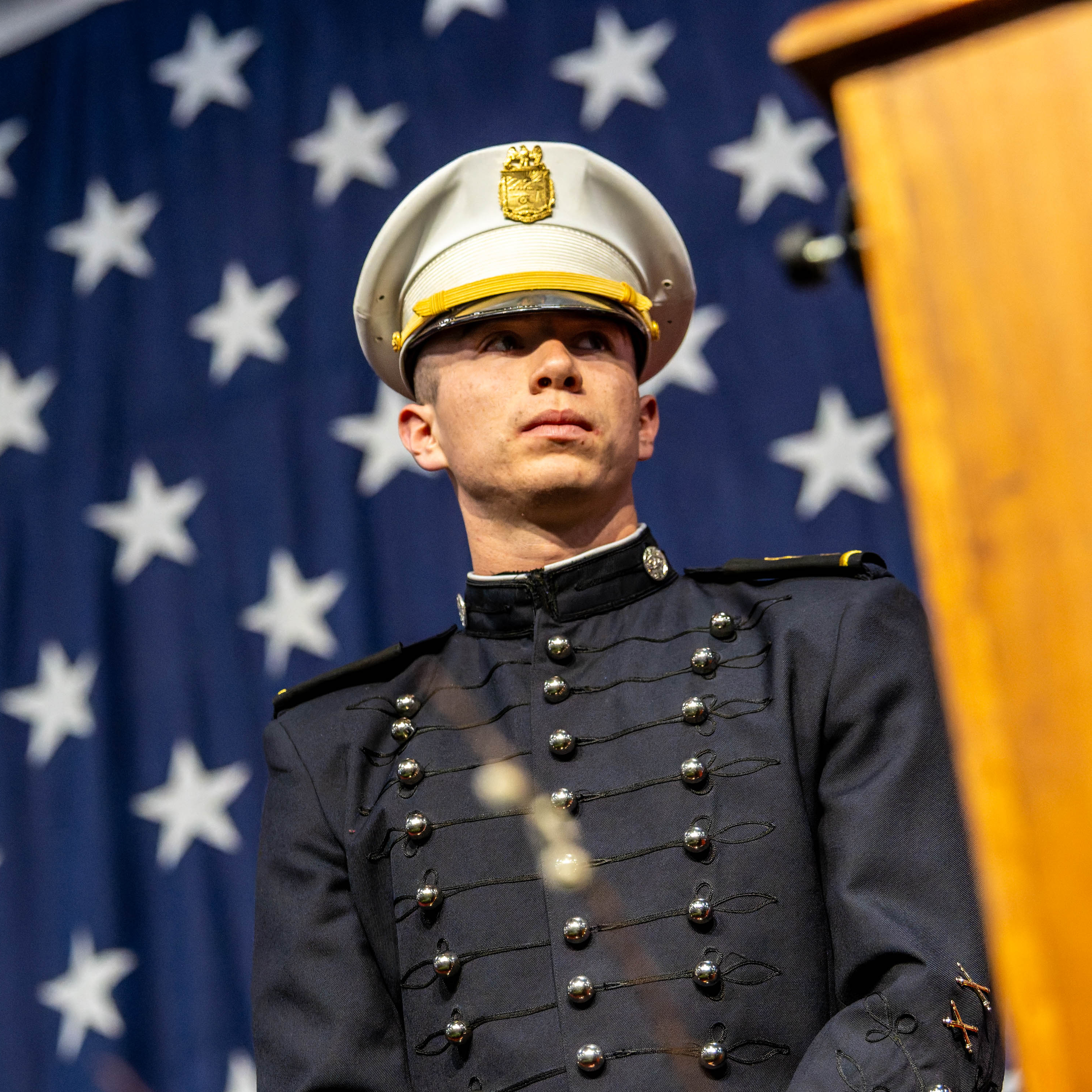 Cadet in dress uniform stands before large American flag during ceremony.