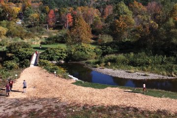 A group of people walk on a woodchip path by a river, surrounded by dense autumn trees; a small footbridge crosses the water in the background.