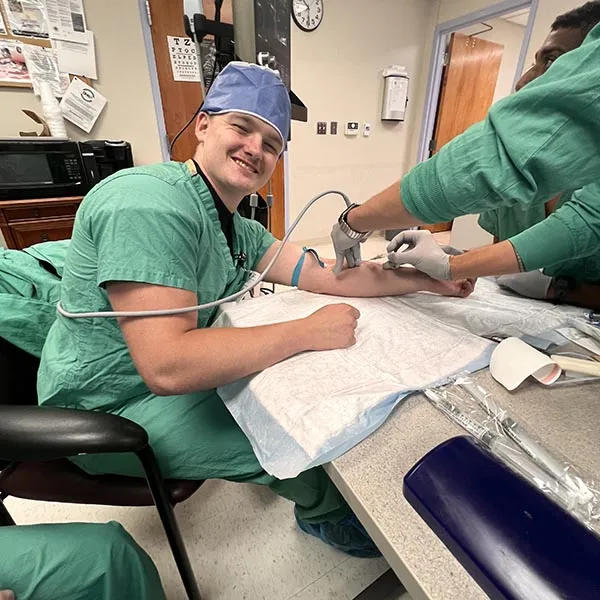 A healthcare worker in green scrubs uses an ultrasound probe and needle on another healthcare worker's arm.